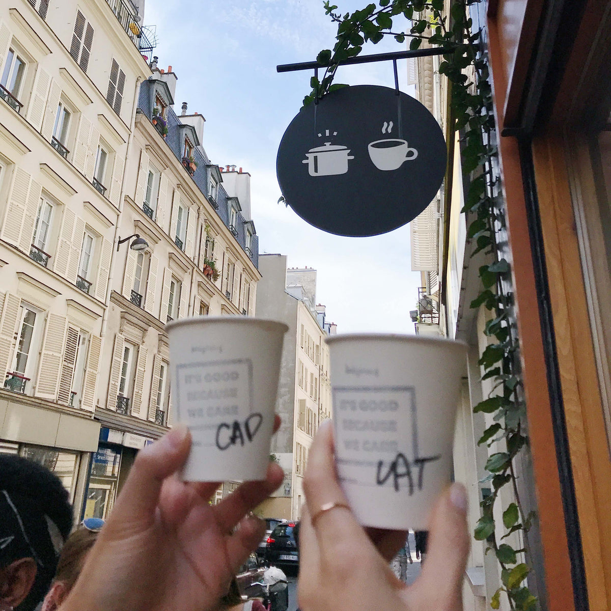 Cheerful toast with coffee cups under a quirky café sign in Paris, embodying the joy of shared moments and good beverages while showcasing a dainty gold band ring by Katie Dean Jewelry.