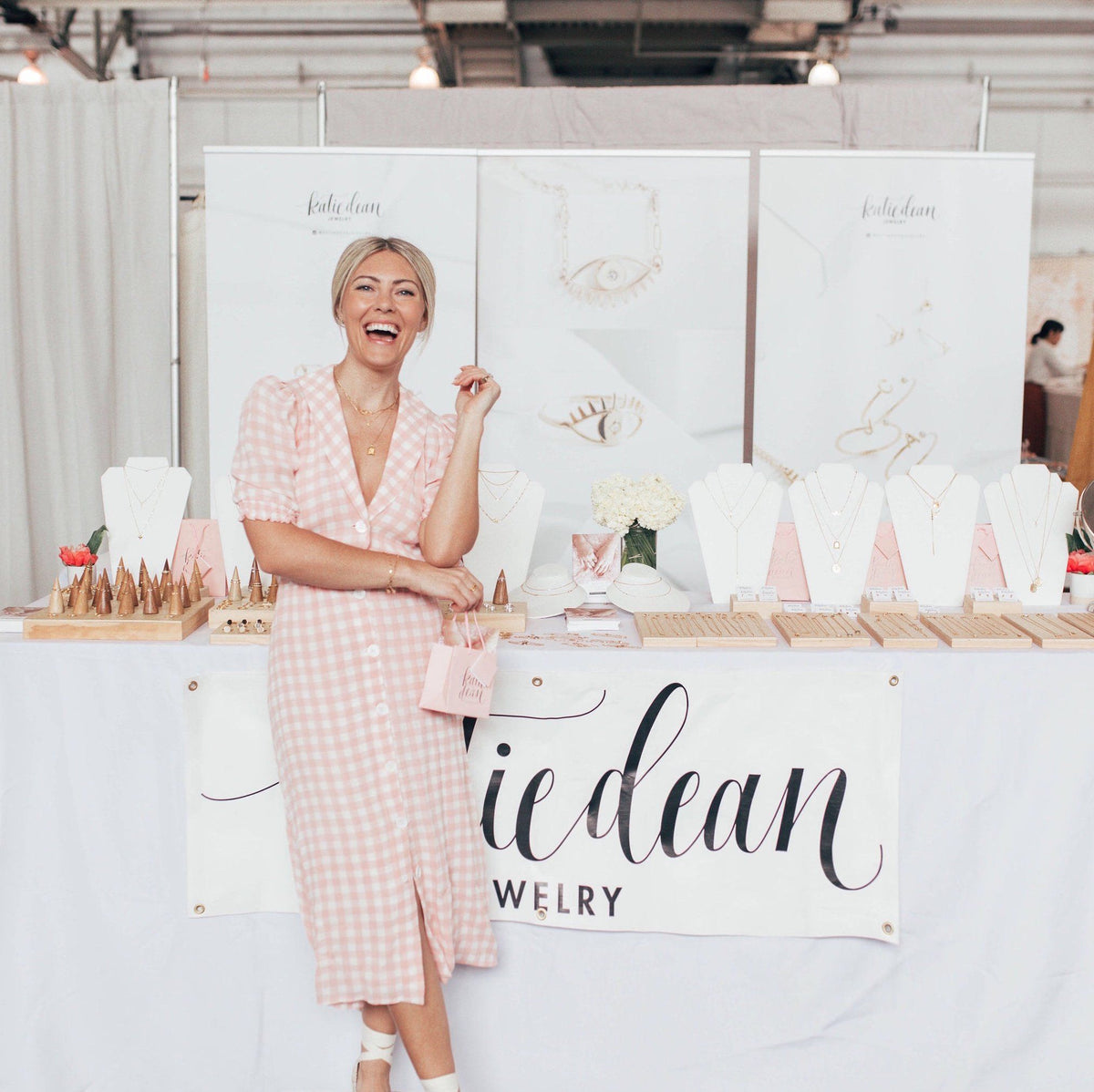 Katie Dean Jewelry in a pink dress standing in front of her jewelry display, a white table with white necklace stands and her dainty gold jewelry laid out.
