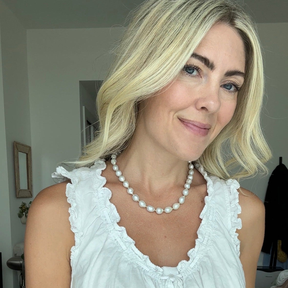 Woman wearing a white sleeveless top with lace details indoors and a pearl necklace.