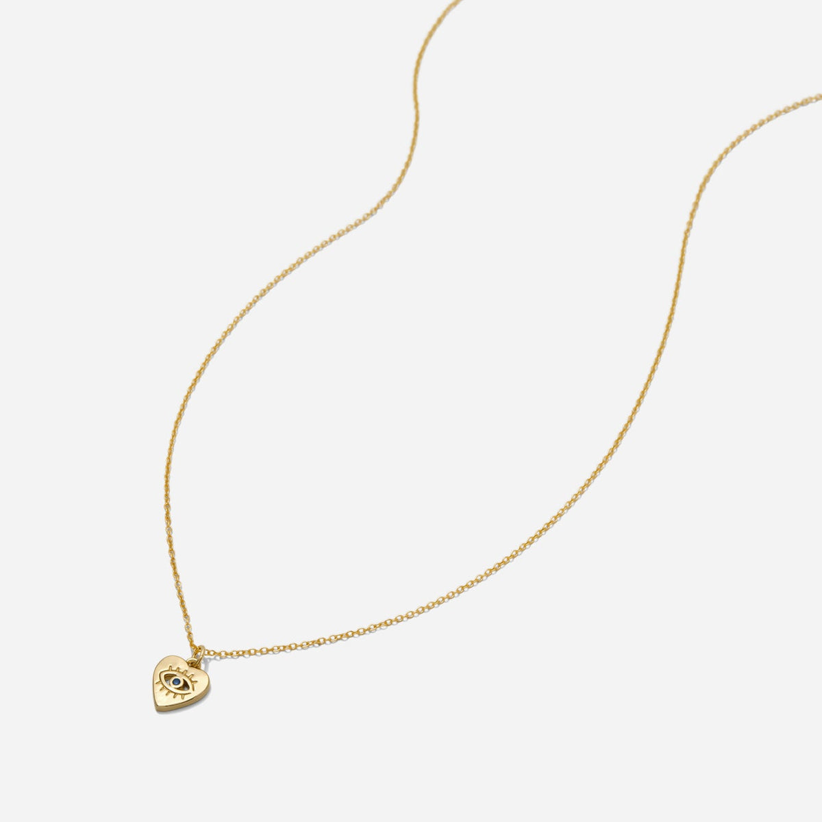 Dainty gold necklace with a heart-shaped pendant with evil eye engraving on a white background, made by Katie Dean Jewelry.