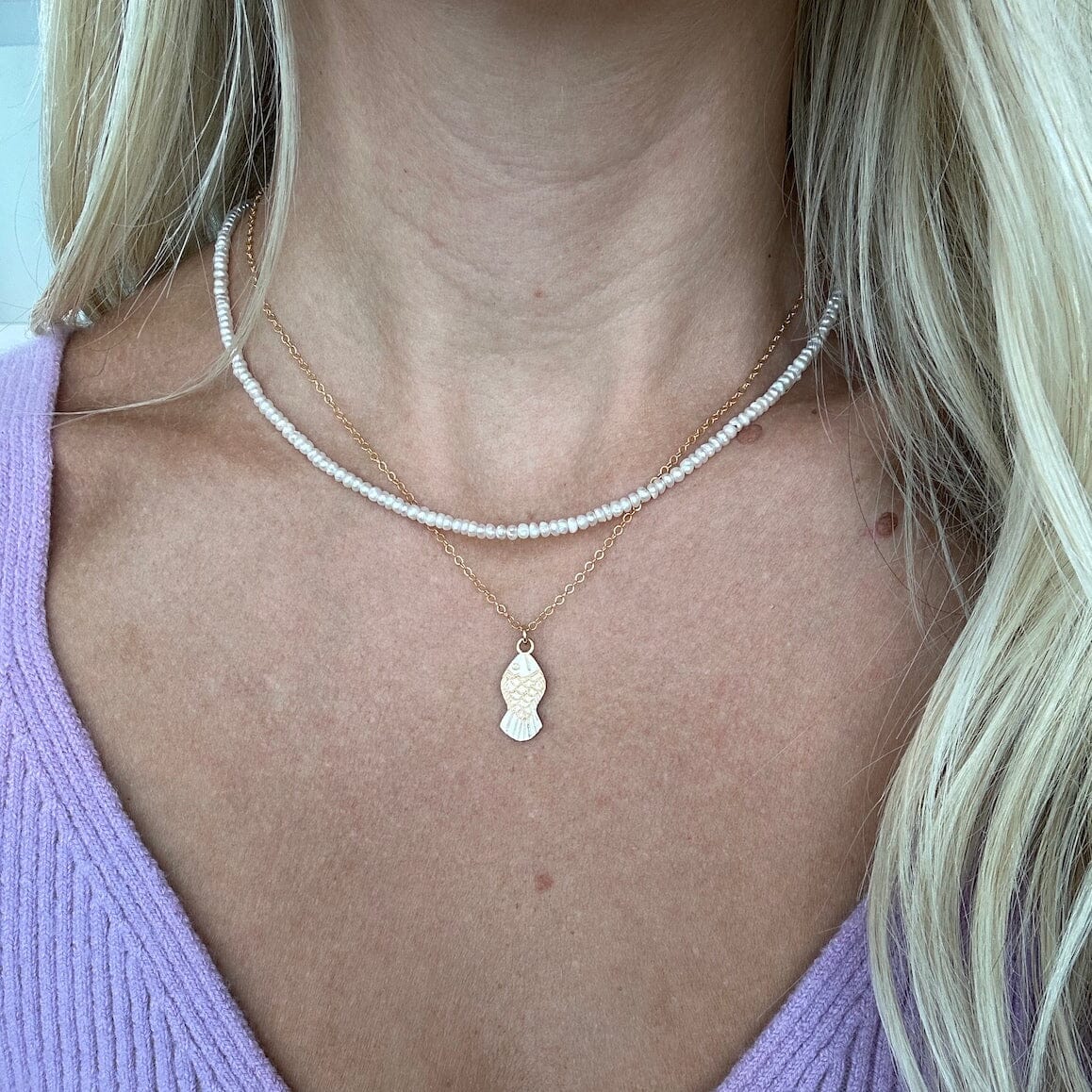 Close-up of a person wearing two necklaces, one small strung pearls and the other a gold chain necklace with a gold fish charm with a purple top.