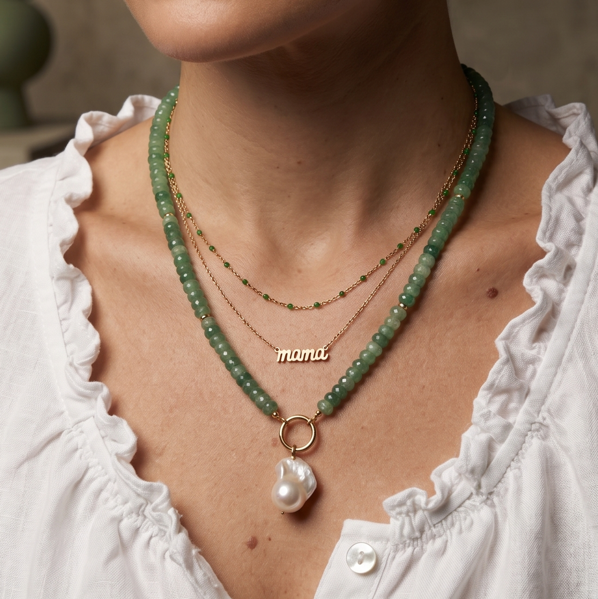 A close-up of a woman wearing a layered necklace set. The set includes a green beaded necklace with a gold ring and a white baroque pearl pendant, a delicate gold chain with a 'mama' nameplate, and a thin gold chain with small green beads.