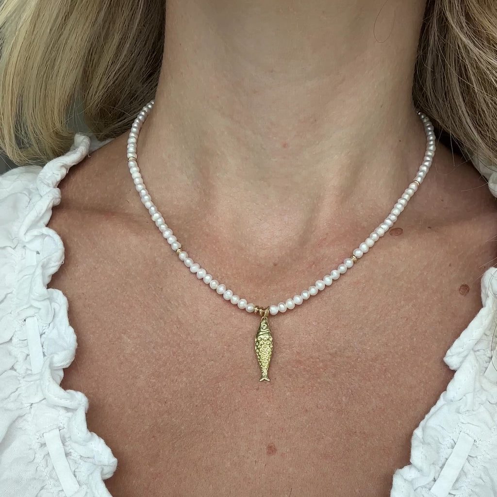Pearl necklace with a gold fish pendant worn by a person in a white blouse.