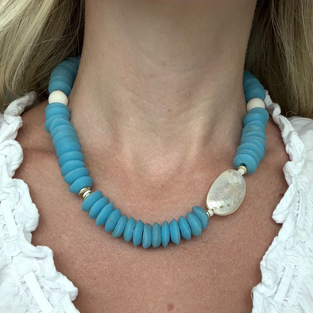 Necklace with blue beads and an abalone shell on a person wearing a white top.